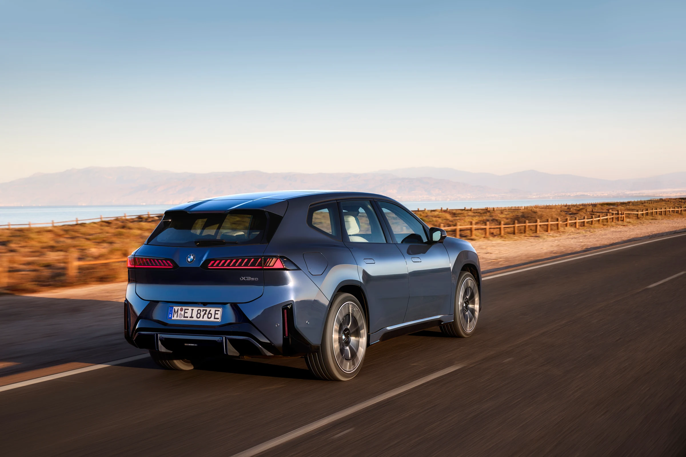 Blauer BMW iX3 50 xDrive der Neuen Klasse in Heck-Dreiviertelansicht auf einer Küstenstraße.